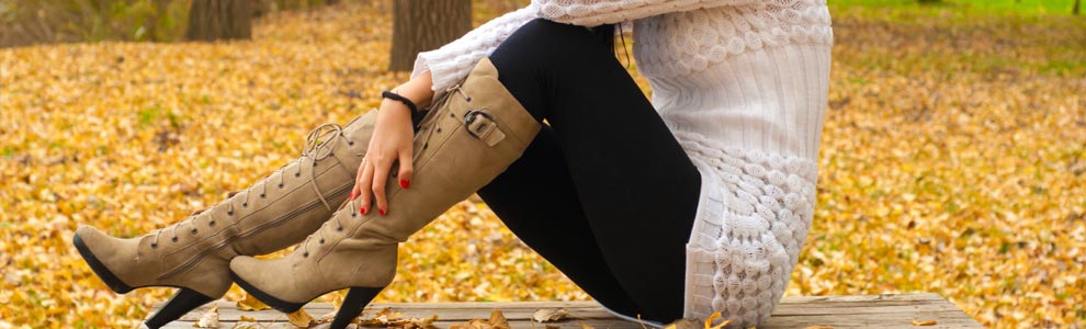 A woman wearing knee high, leather, high heeled boots sits on an autumn park bench